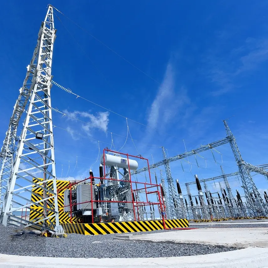 A power transformer with red and white casing is secured within a high-voltage electrical substation, surrounded by metallic structures and power lines against a clear blue sky.