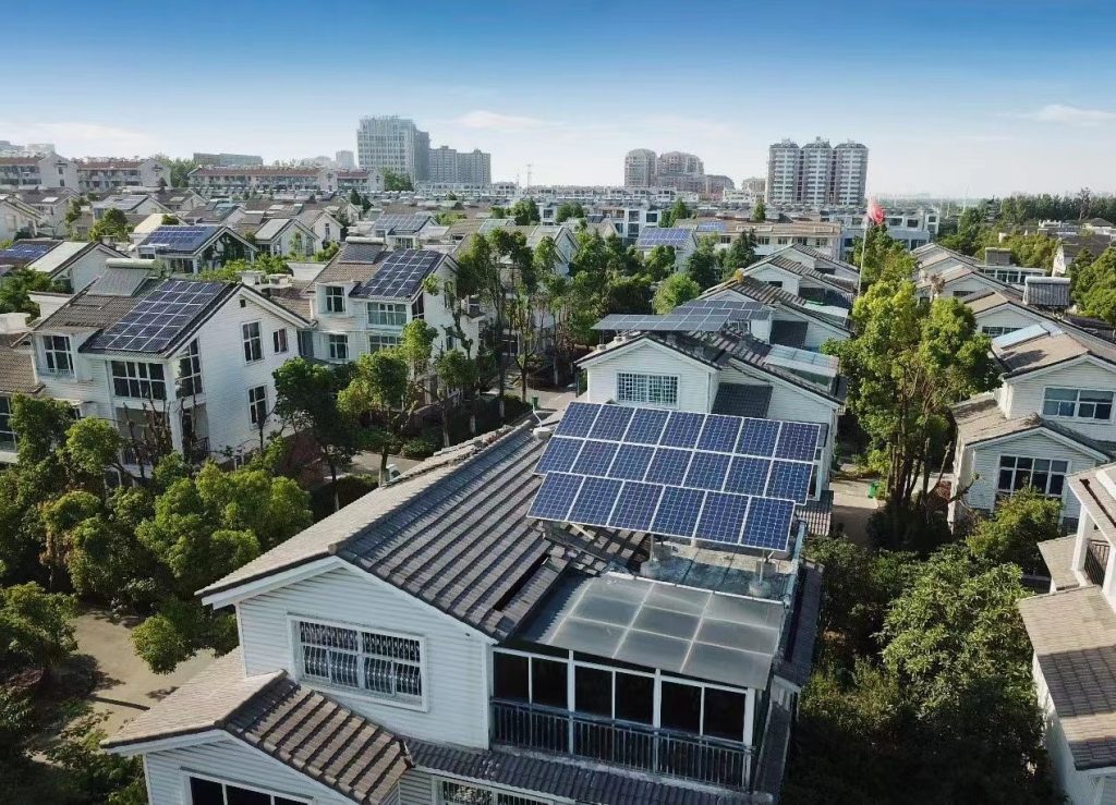 This image displays a suburban area where several houses are equipped with solar panels on their roofs. The neighborhood is lush with greenery, consisting of numerous trees interspersed between the homes. The architecture of the houses appears modern with a mix of gabled and flat roofs, some of which are entirely covered with solar panels, suggesting a community committed to sustainable energy use. In the background, multi-story buildings can be seen, indicating the proximity of a more urban environment. The sky is clear and blue, indicating good weather conditions for solar energy production.